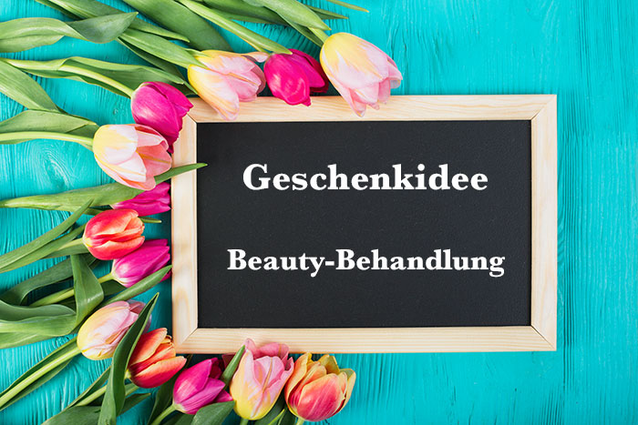 Schiefertafel mit Aufschrift "Geschenkidee Beauty-Behandlung" auf türkisem Hintergrund und umrahmt von rosefarbenen Tulpen